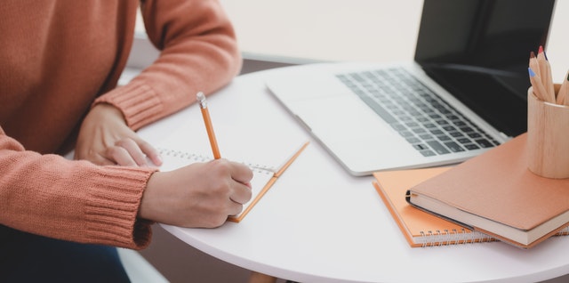 Person writing on a desk with a computer and books. 